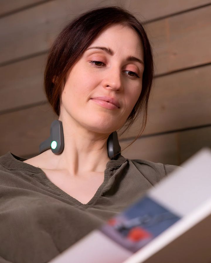 Woman reading a book with the Pulsetto Lite worn at the base of her neck, its green status light on